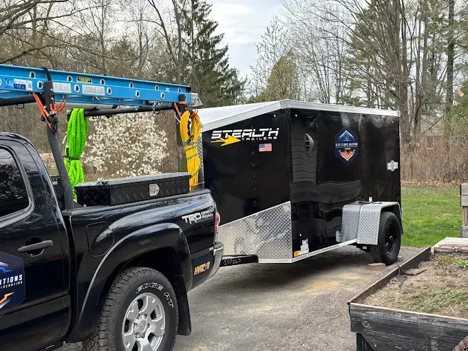 Black truck towing a black enclosed trailer, a ladder on top. Exterior setting.