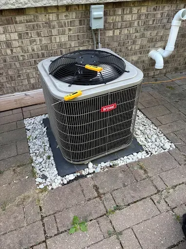 Bryant air conditioner on a brick patio with rocks, near a brick wall and white pipe.