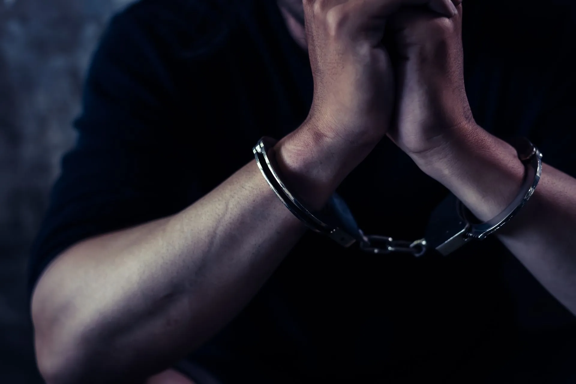 Man's hands clasped in handcuffs, close-up, dark setting.