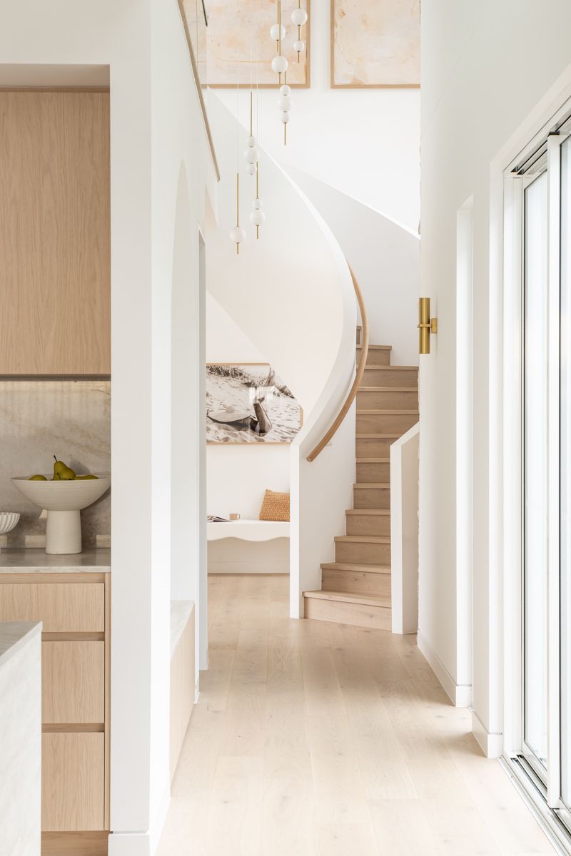 Hallway with wooden stairs, light wood floors, and white walls. A kitchen area with wood cabinets is visible on the left. — SDC Building Design In Kiama, NSW