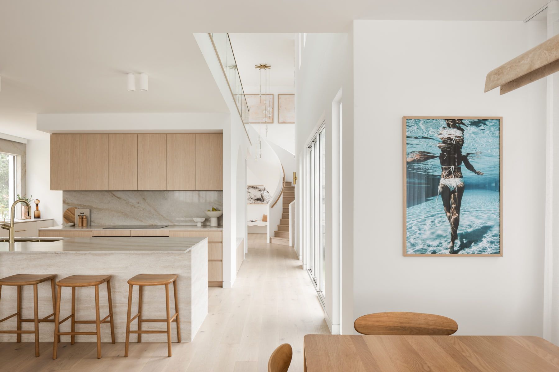 Modern, light-filled kitchen with light wood cabinets, marble counters, and a hallway with a staircase and artwork — SDC Building Design In Kiama, NSW