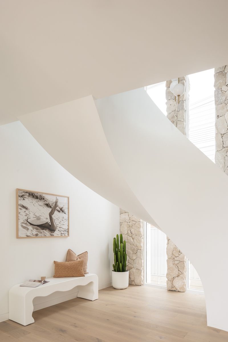 White interior with curved staircase, art, bench, cactus, and stone column. — SDC Building Design In Kiama, NSW