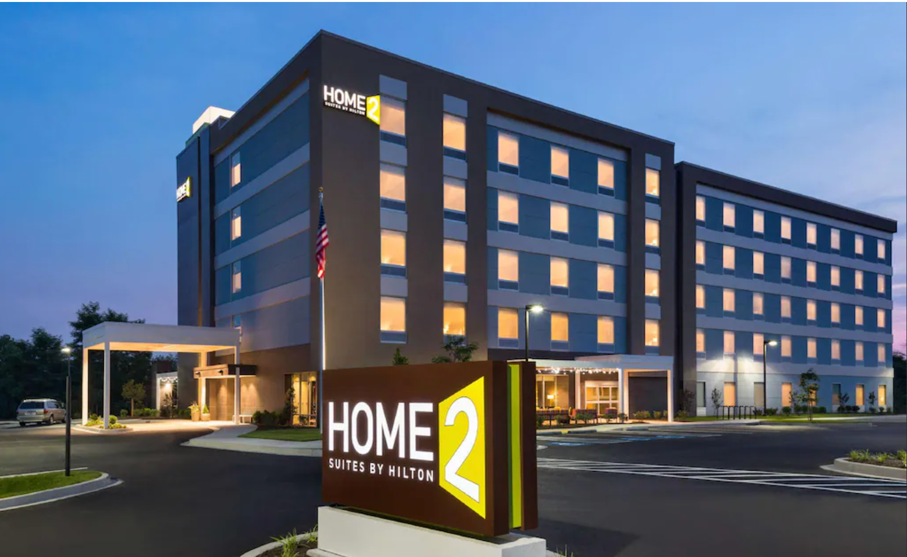Home2 Suites hotel at dusk, exterior view; brown building, lit windows, signage, and entrance.