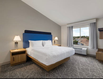 Hotel room with a bed, nightstands, and a window with gray curtains; the wall is gray, the headboard is blue.