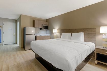 Hotel room with a queen-sized bed, mini-kitchen, refrigerator, and bedside table with lamp.