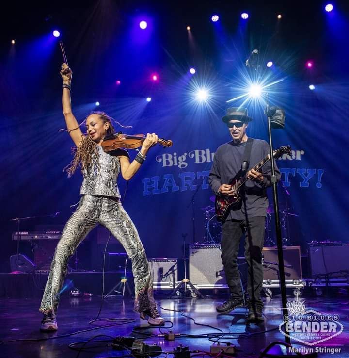 A women holding a violin fiddle and bow in left hand with a man wearing hat and dark sunglasses stands beside wih arms crossed in front of his body