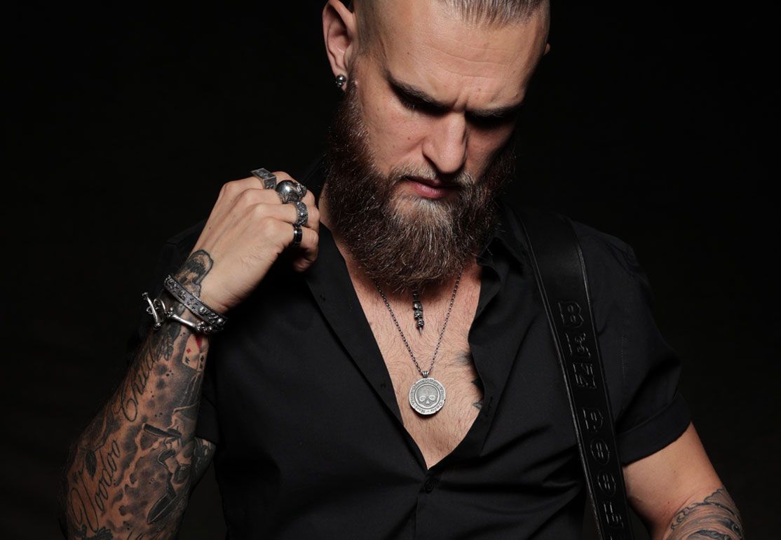 A man with a beard and tattoos is wearing a black shirt and a necklace.