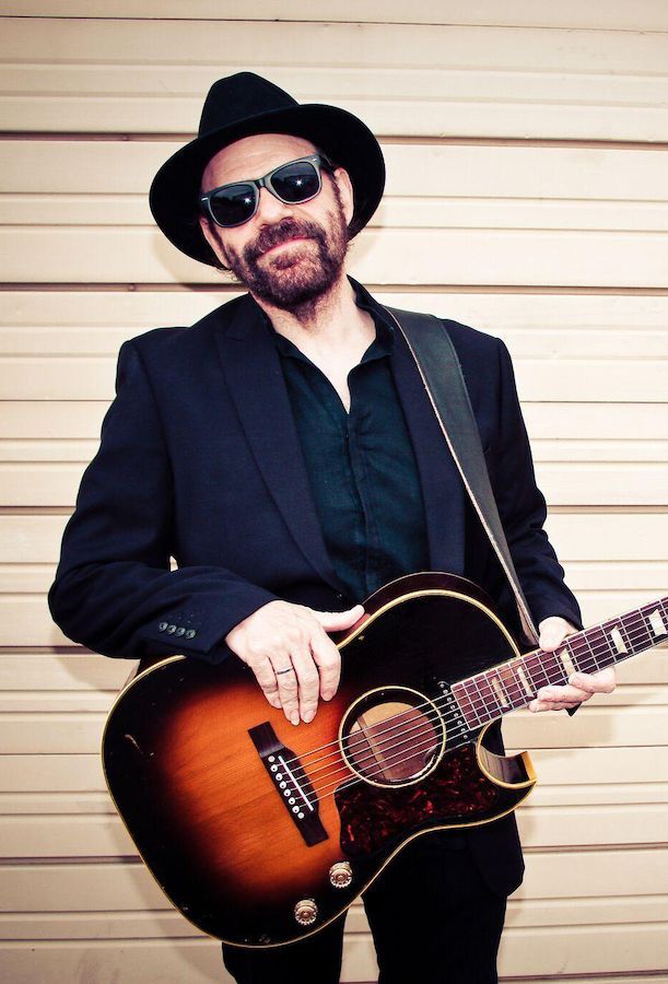 Colin Linden wearing sunglasses and hat while smiling and holding guitar smiling
