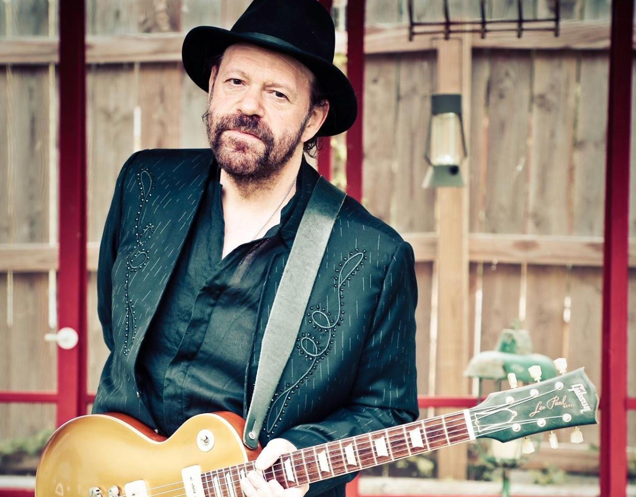 Man in black jacket and hat, holding a gold guitar, standing in front of a red structure.