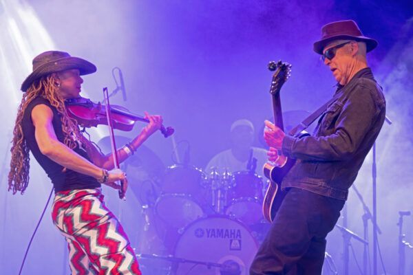 A band of seven members posing together, instruments visible. Indoor studio setting with windows and vintage televisions.
