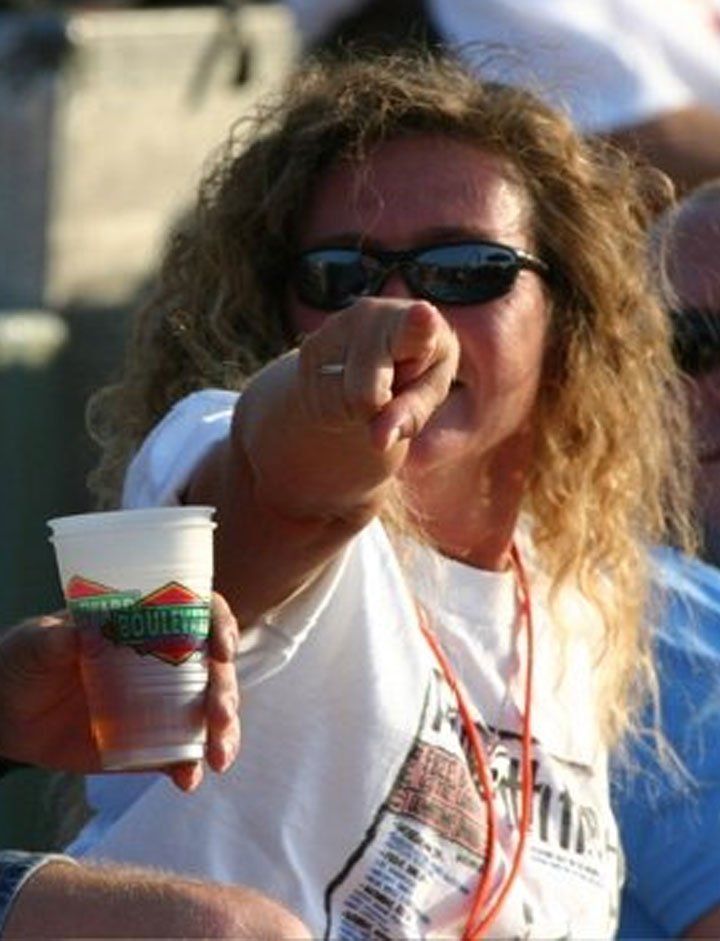 A woman wearing sunglasses is pointing at someone while holding a cup that says ' harley davidson ' on it
