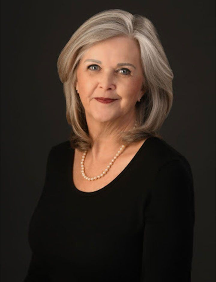 A woman with gray hair is wearing a black shirt and a pearl necklace.