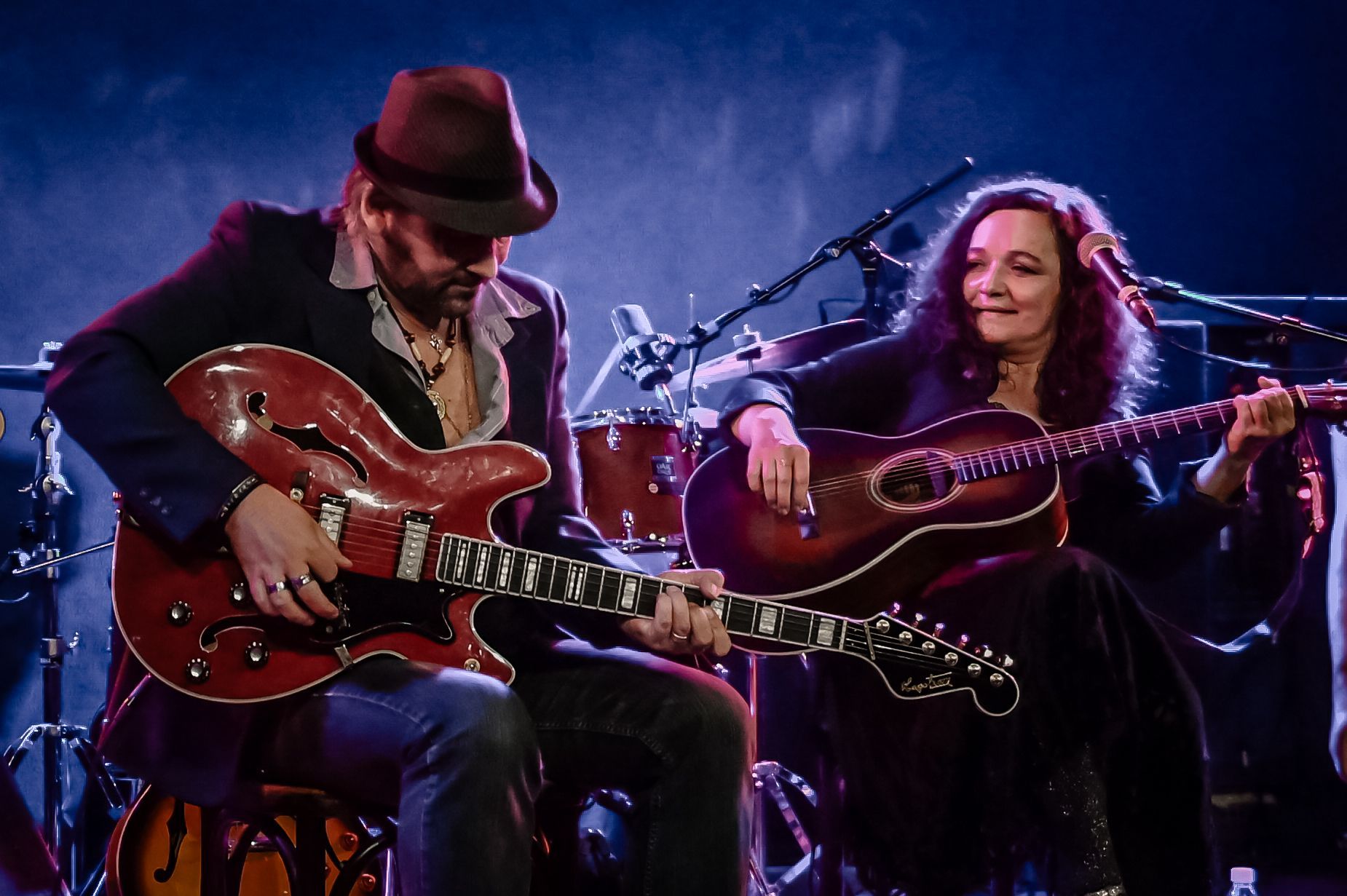 Woman singing into a microphone, man playing guitar. Stage lighting, dark background.