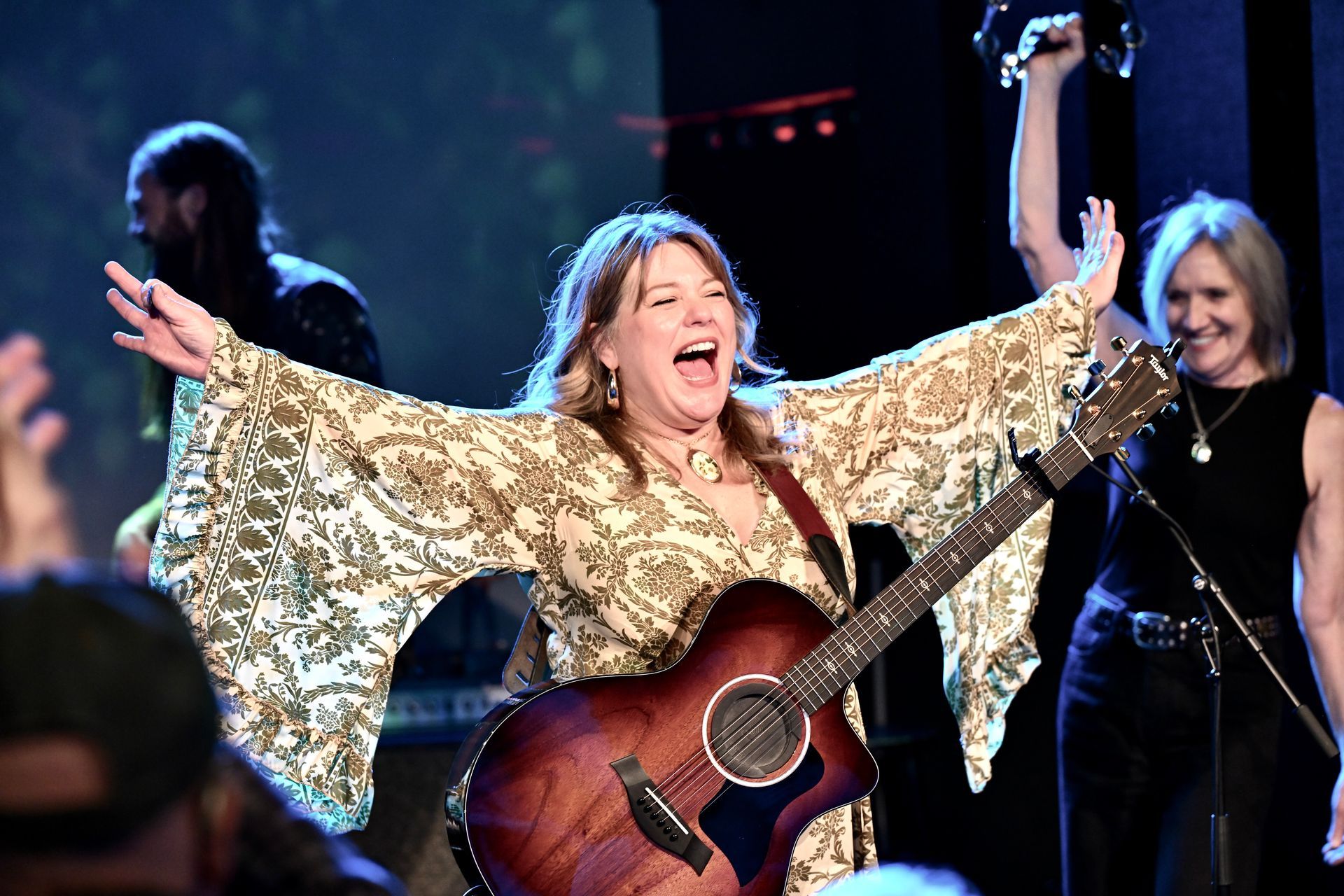 Woman in coral dress plays a sparkly blue guitar on stage, singing into a microphone.