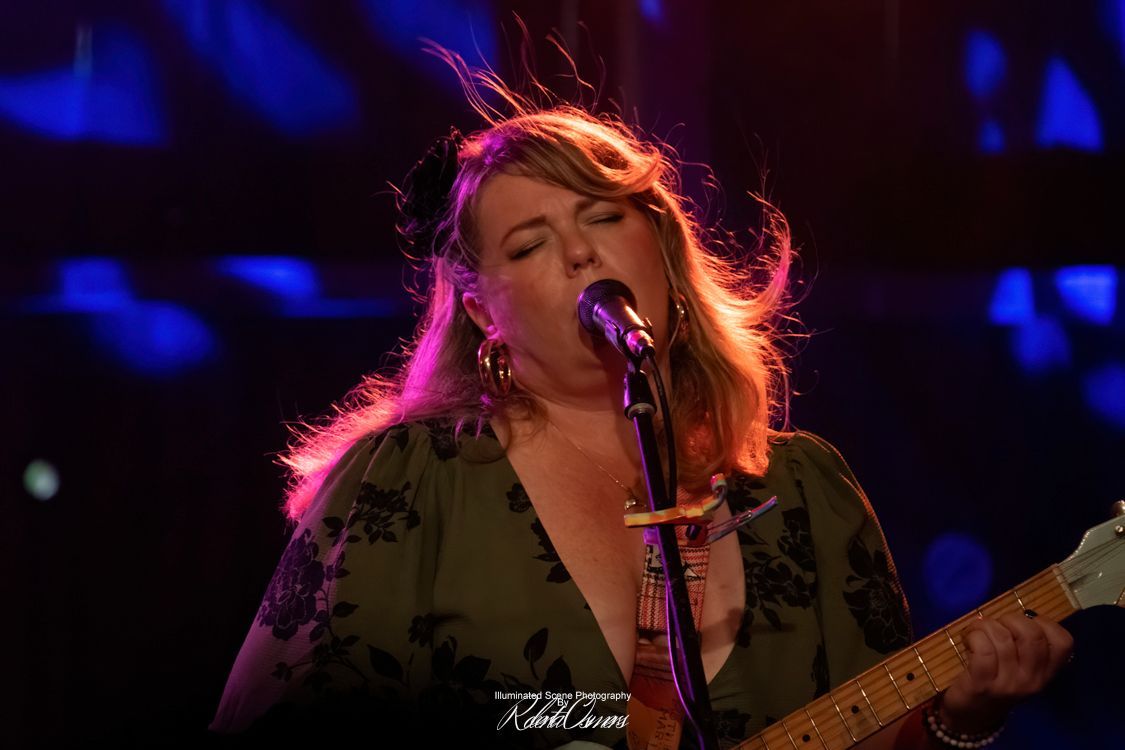 Woman in coral dress plays a sparkly blue guitar on stage, singing into a microphone.