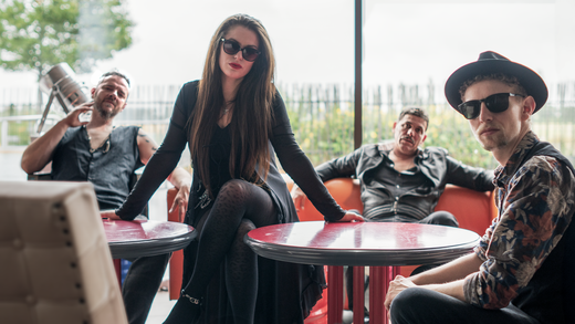 Band in sunglasses poses in a cafe; a woman leans on a table, others seated.