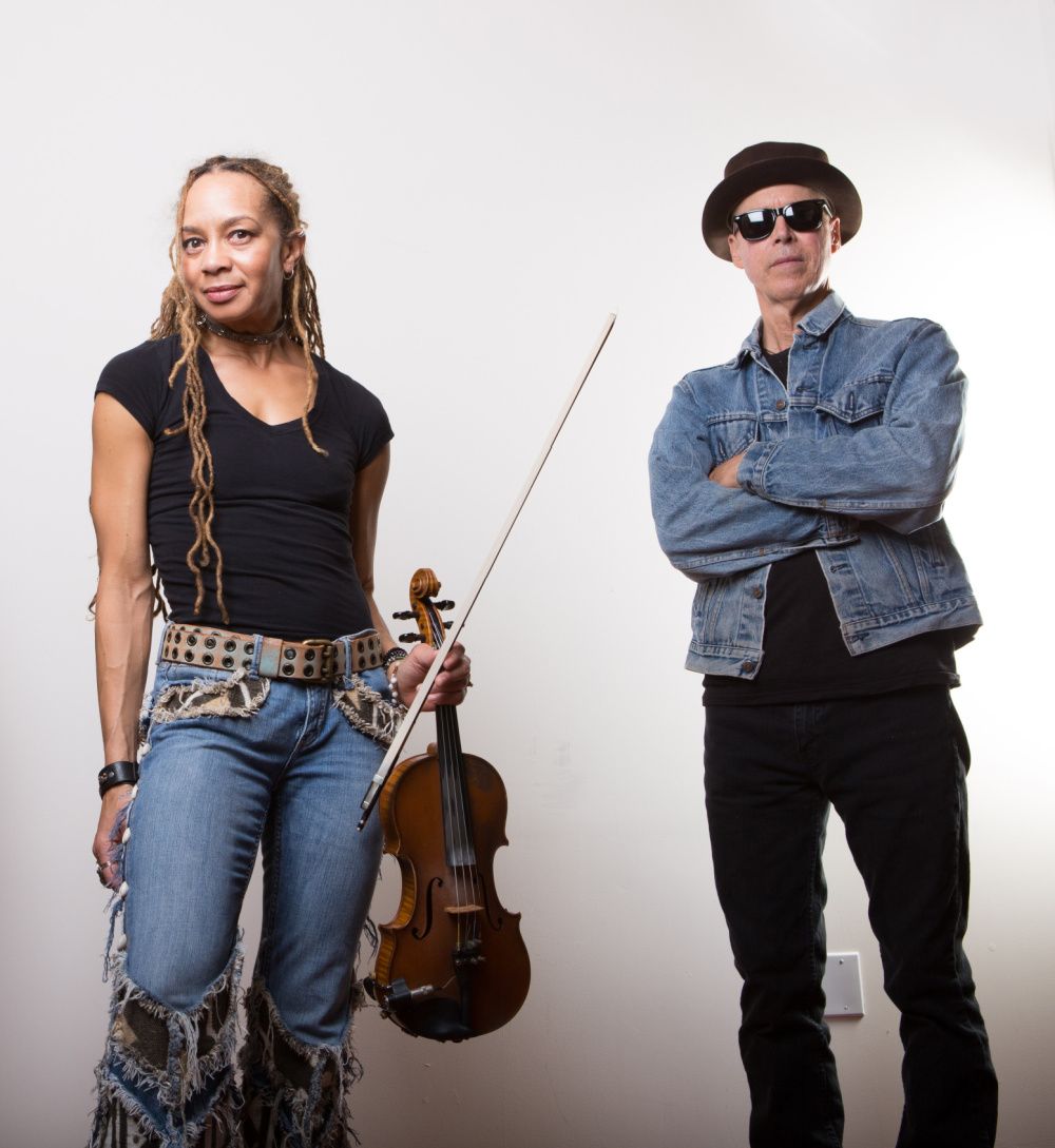 A women holding a violin fiddle and bow in left hand with a man wearing hat and dark sunglasses stands beside wih arms crossed in front of his body