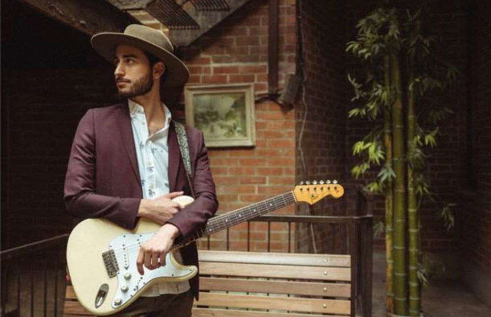 A man in a hat is holding a guitar in front of a brick wall.