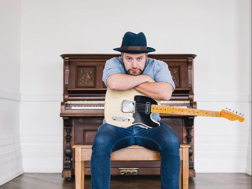 Steve Marriner sitting on a piano bench leaning on guitar sitting on his thighs 