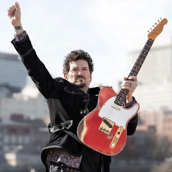 A man in a black jacket is holding a red electric guitar