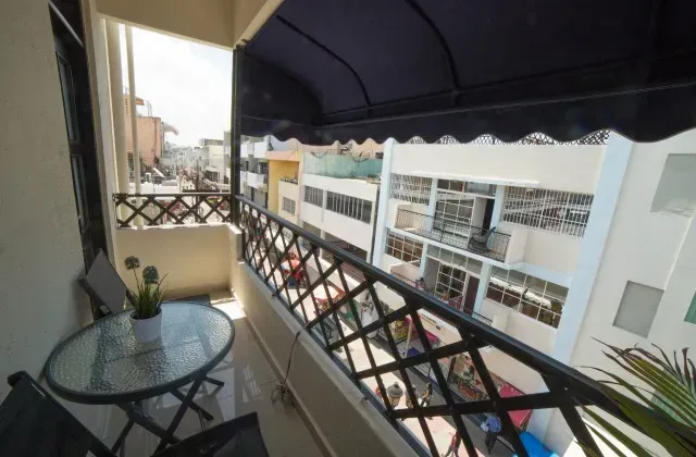Balcony overlooking a street.  Black wrought iron railing, table, chairs. Buildings in background. Blue awning.
