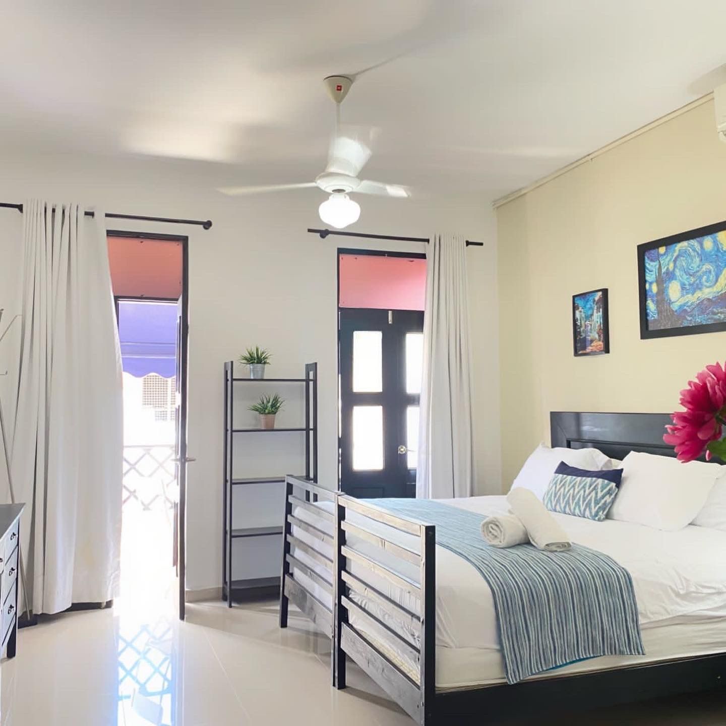 Bedroom with white bedding, blue accents, black bed frame, open balcony doors, and a ceiling fan.