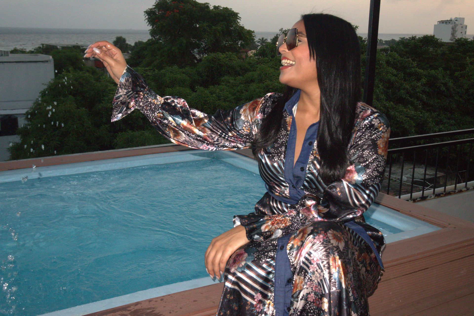Woman in metallic robe, sunglasses, near pool, gesturing with trees and ocean in the background.