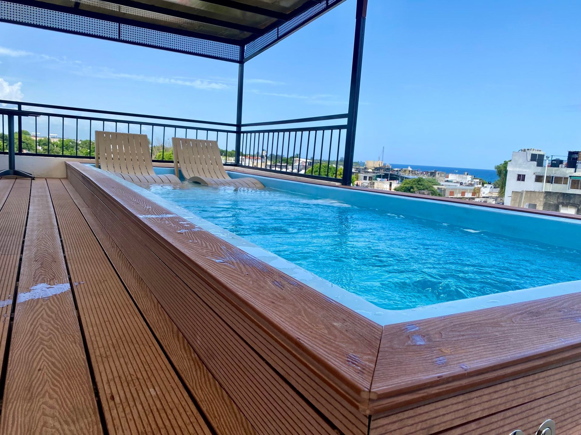 Rooftop pool with wood decking, two lounge chairs, and a partial view of buildings and ocean under a blue sky.