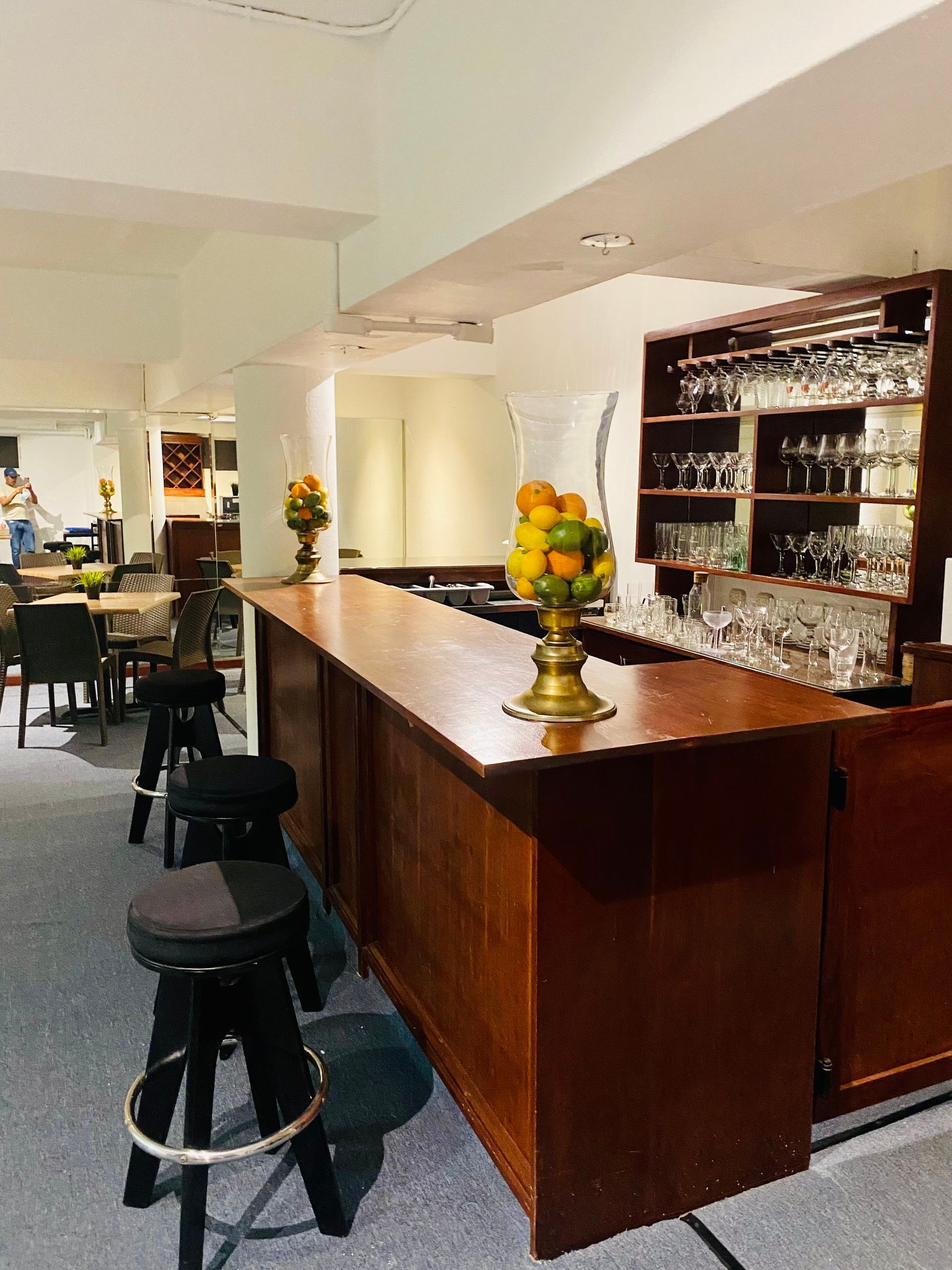 Wooden bar with stools, shelves of glassware, and fruit in a bowl.