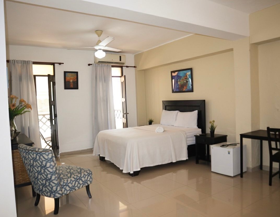 Hotel room with bed, chair, desk, and balcony access. White and beige tones.