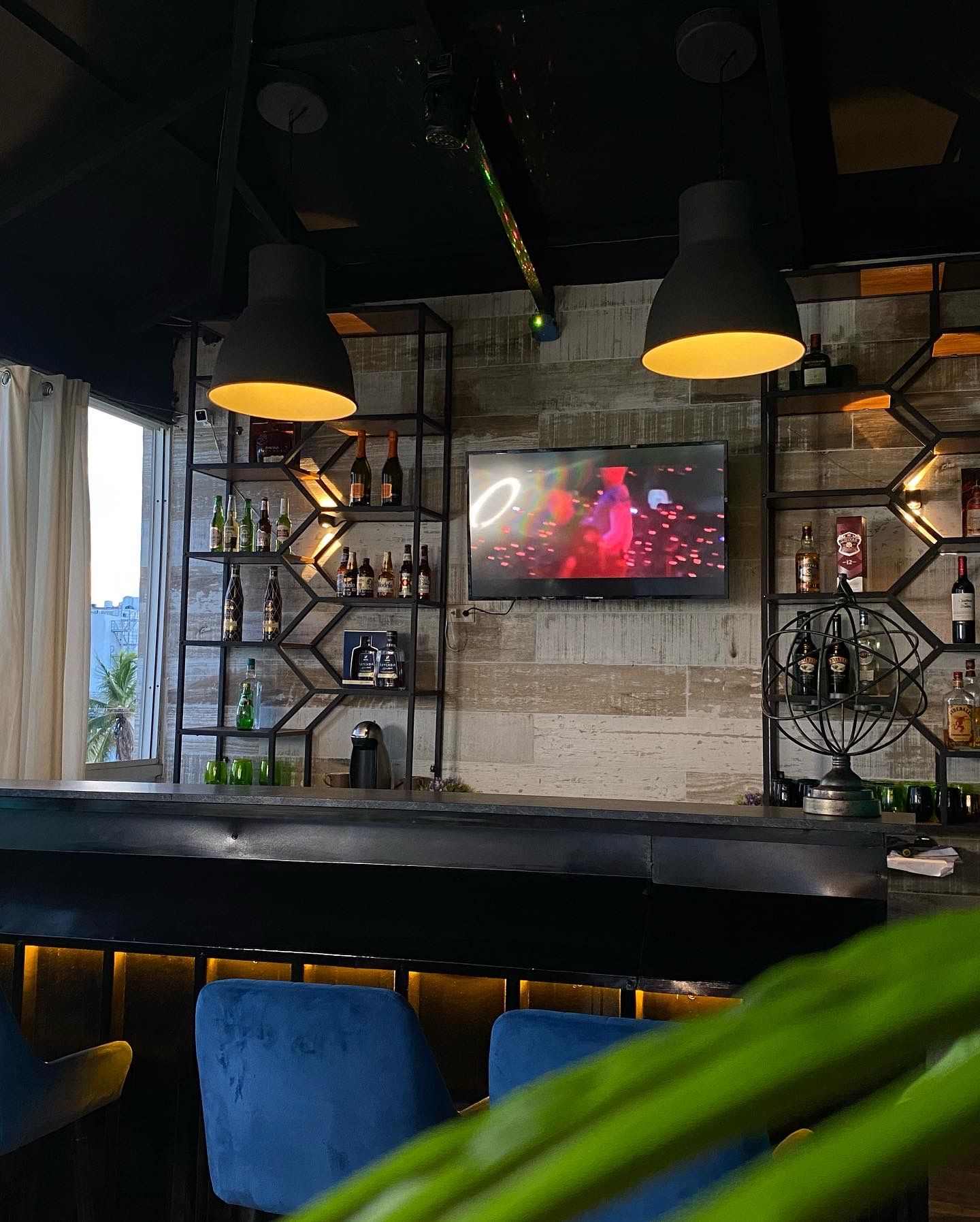 Interior of a bar with shelves of liquor, a TV, pendant lights, and blue bar stools.