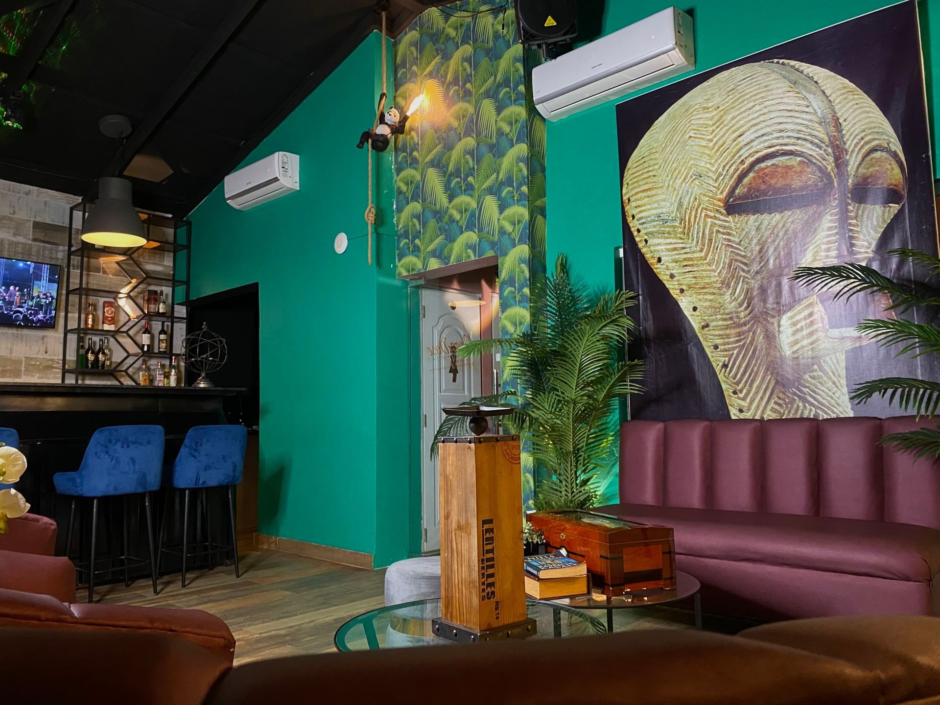 Interior of a bar with teal walls, a dark ceiling, and a large tribal mask artwork; includes seating and bar area.