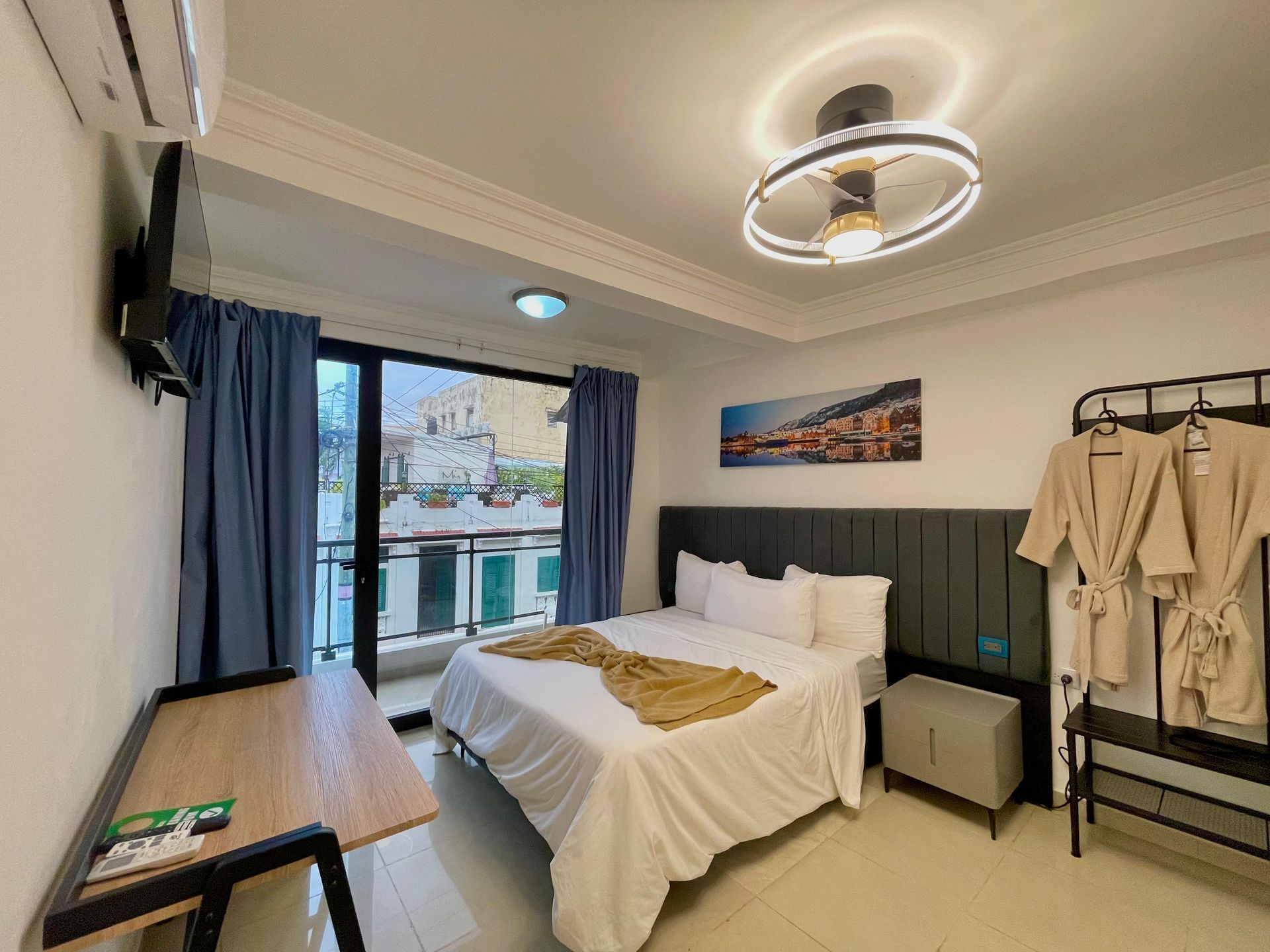 Hotel bedroom with a bed, desk, balcony, and robe rack; blue and beige color scheme.
