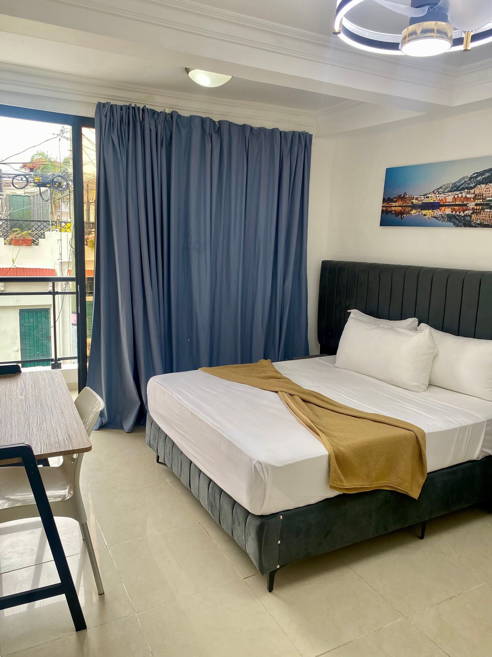 Bedroom with bed, blue curtains, and small balcony. Gold blanket on the bed, table on the left.