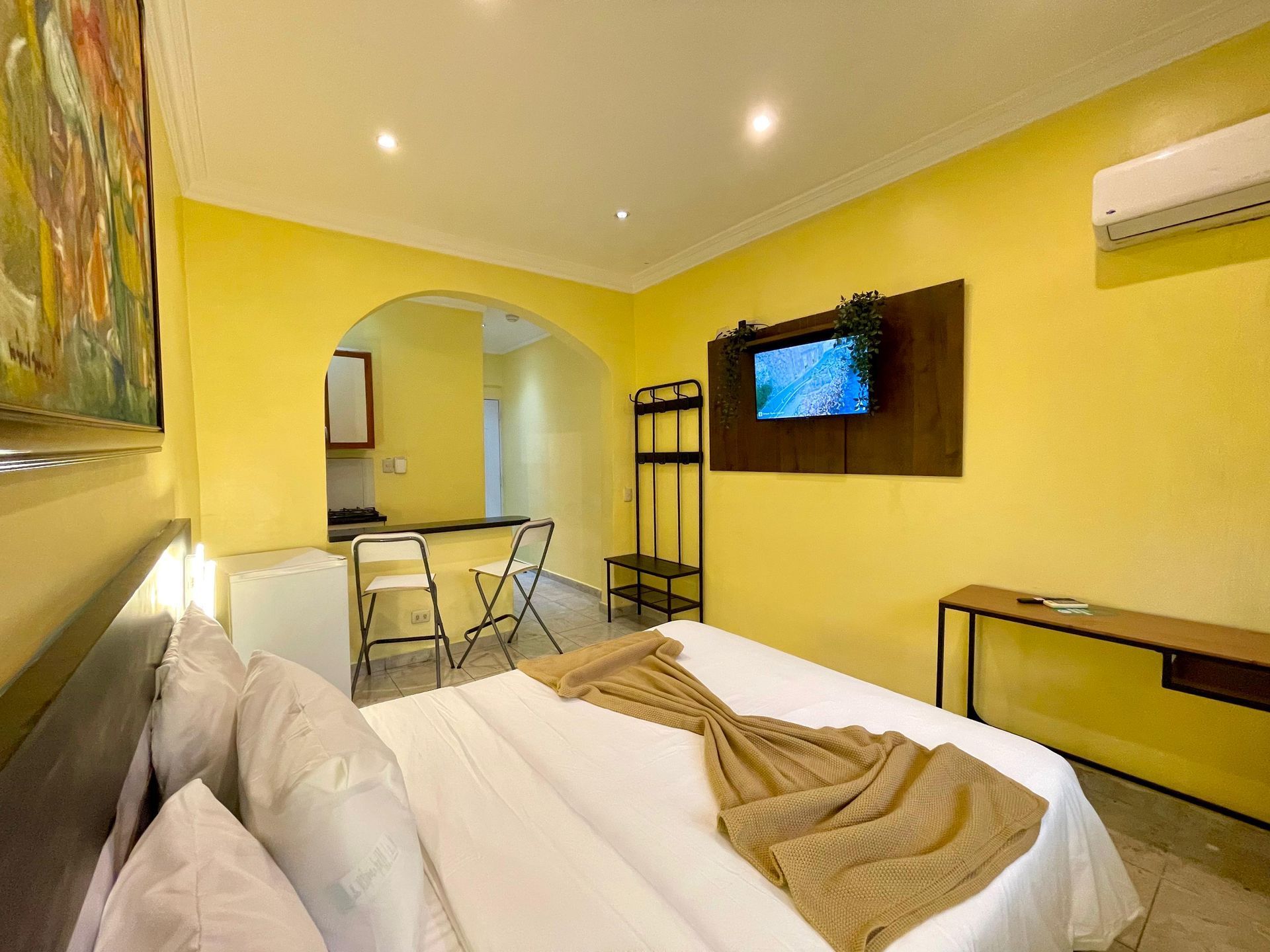 Yellow hotel room with bed, TV, mini-fridge, and a small kitchenette.