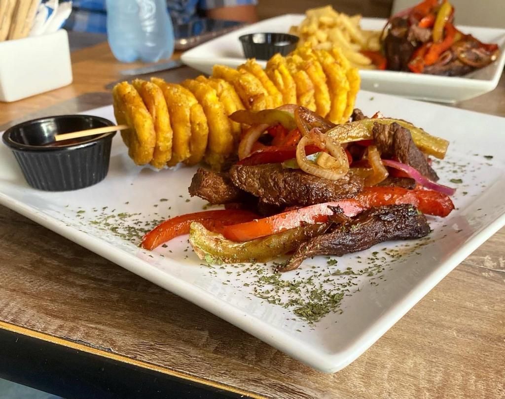 Plate of grilled steak with peppers, spiral fried potatoes, dipping sauce, and side dish of french fries.