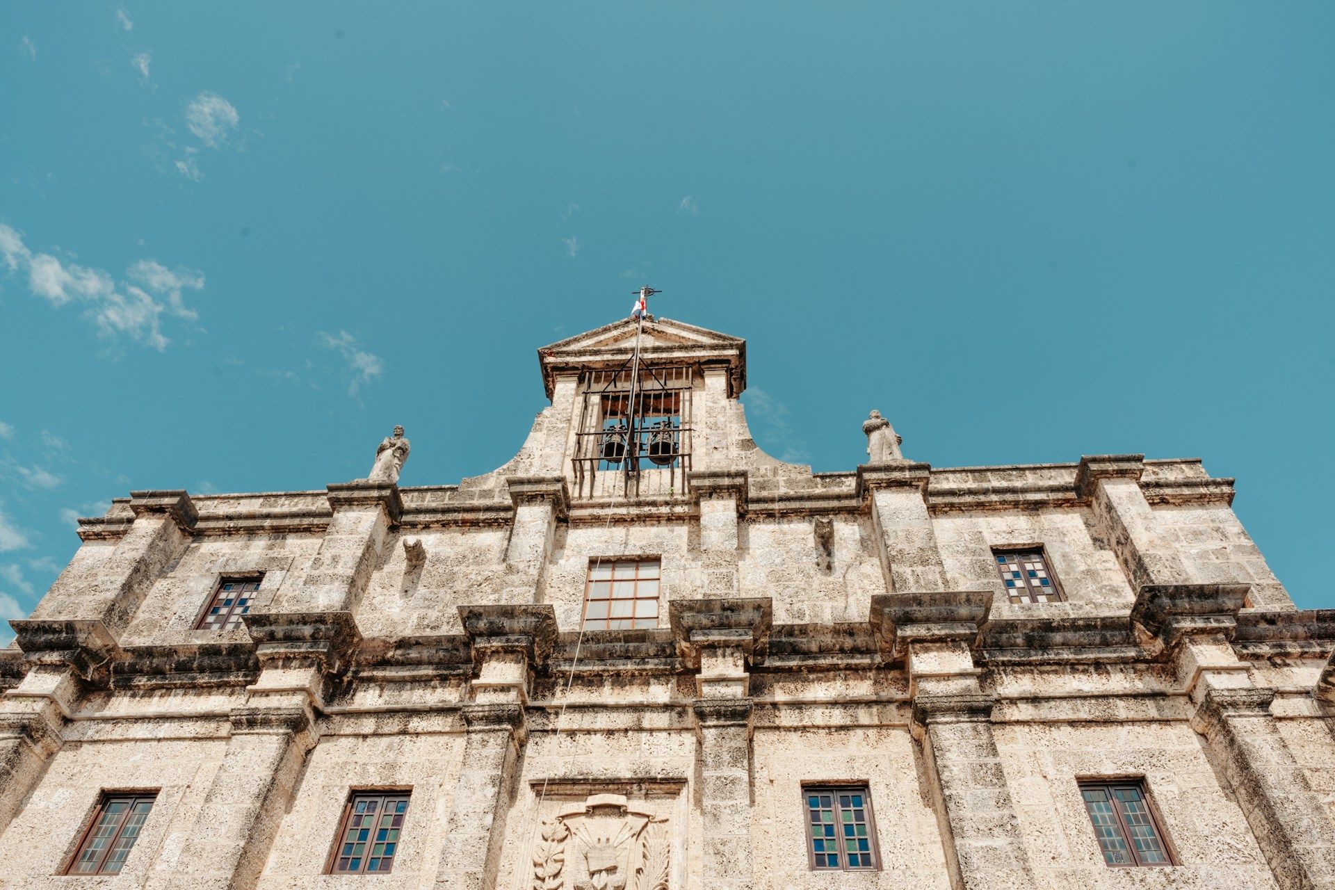 Low-angle view of a beige stone building with windows and a bell tower against a bright blue sky.