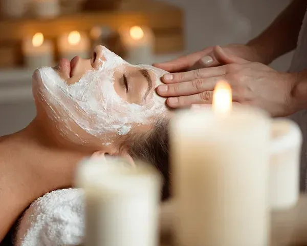Woman with facial mask receiving massage, lit by candles.