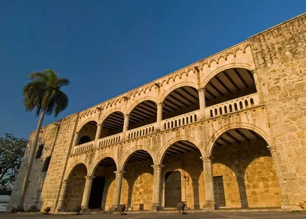 Two-story stone building with arched walkways, tall palm tree, against a blue sky.