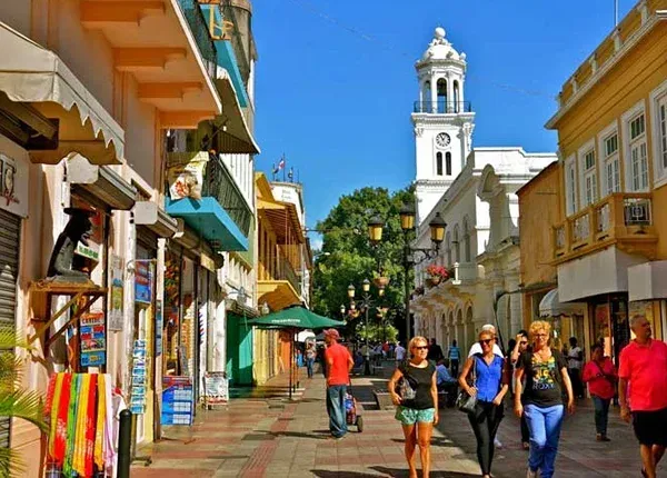 A pedestrian street in a city, lined with colorful buildings and people walking. A church tower is in the background.