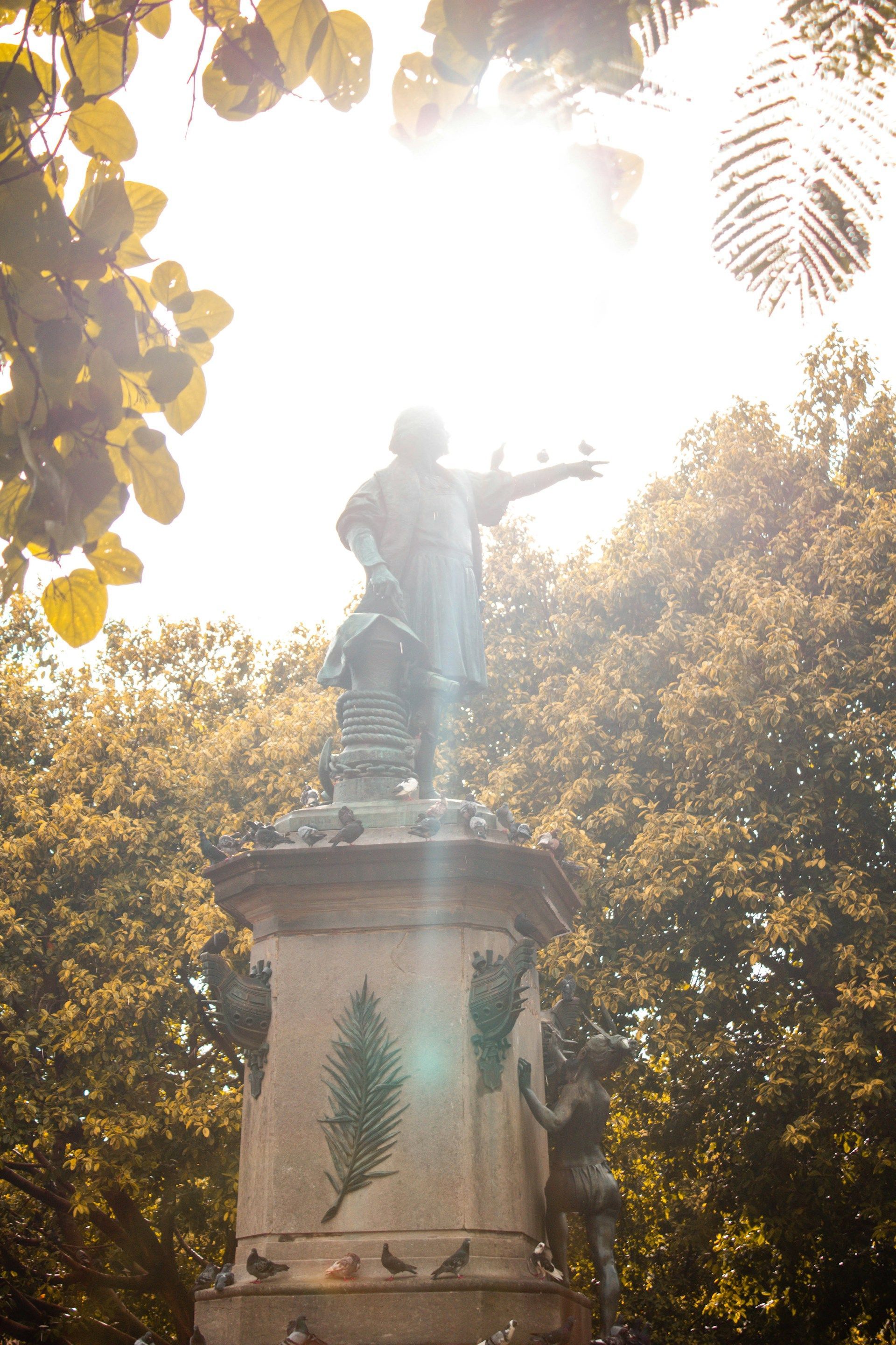 Statue of a man atop a pedestal in a park. Sunlight streams through trees.