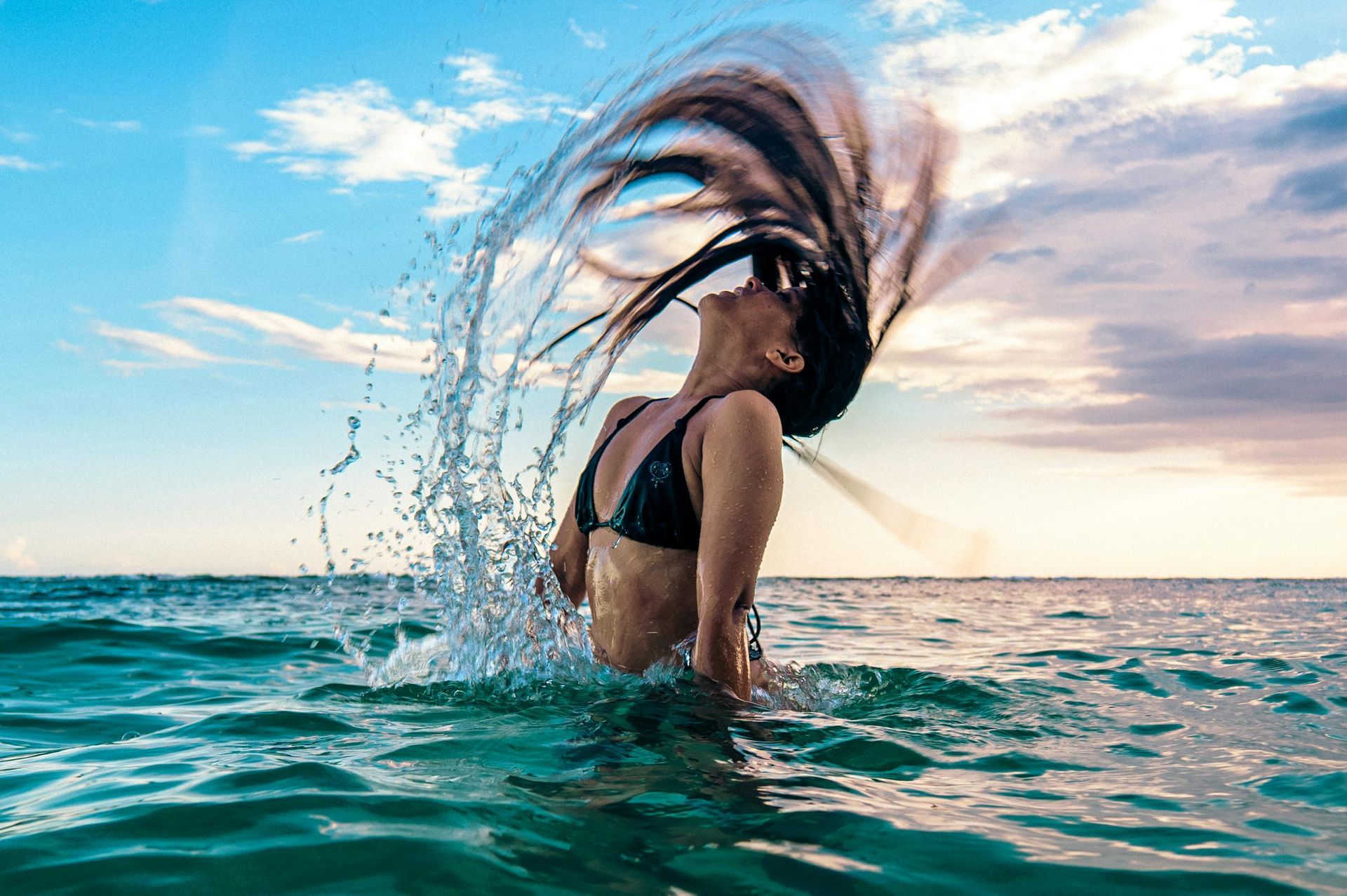 Woman in black bikini tossing wet hair back in ocean, blue sky.
