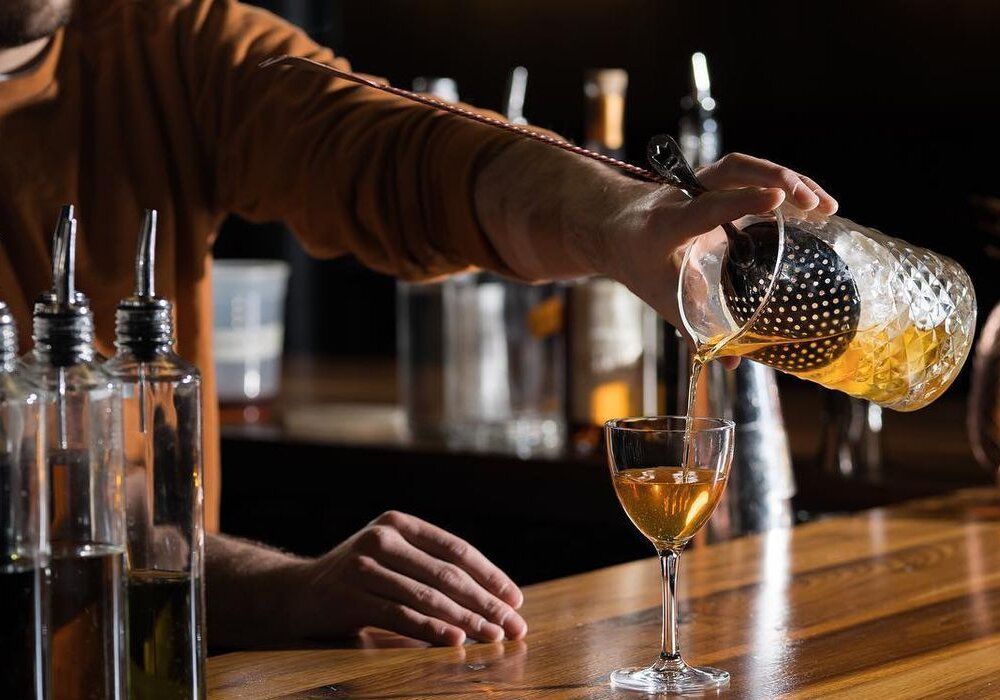 Bartender pours a golden cocktail from a strainer into a stem glass at a bar.