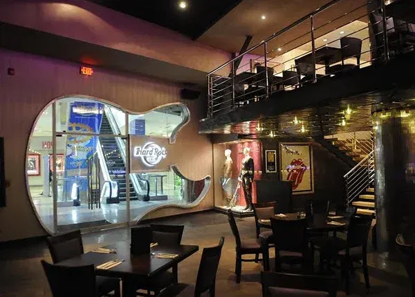 Interior of a Hard Rock Cafe. Guitar-shaped window shows entrance. Tables, chairs, and mezzanine seating area visible.