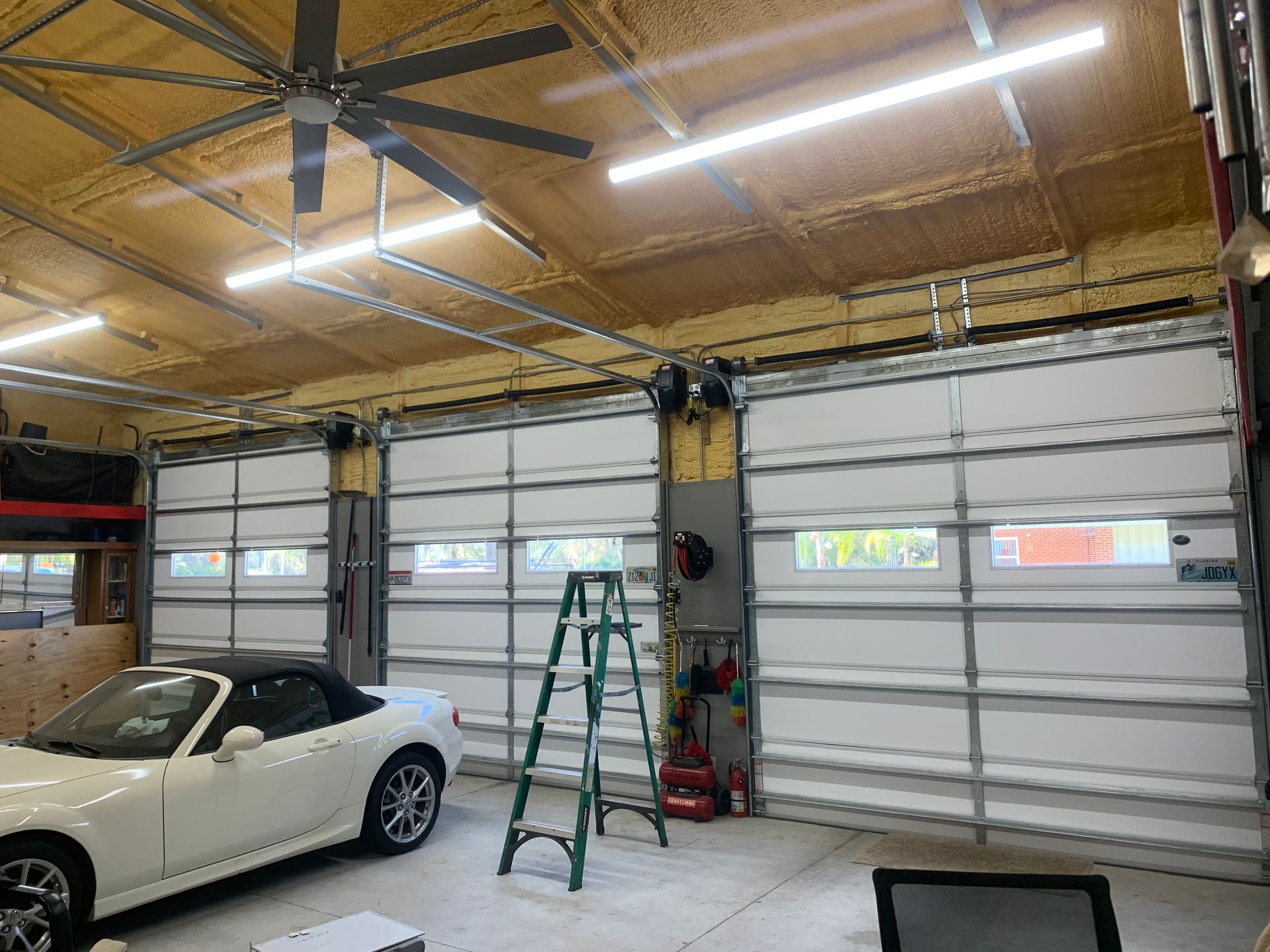 A white car is parked in a garage with a ceiling fan and a ladder.
