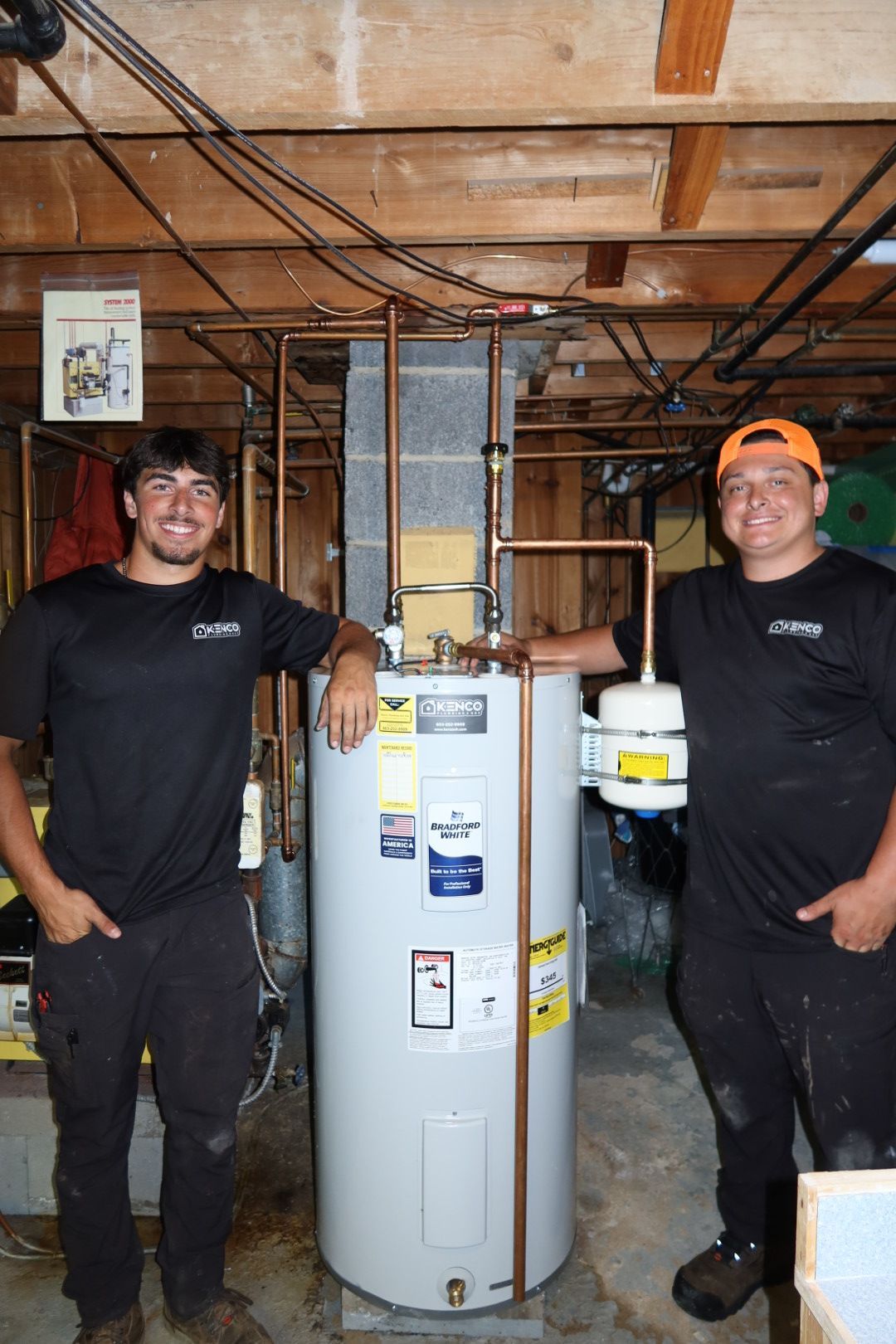 Two men stand proudly beside a new water heater in a basement.