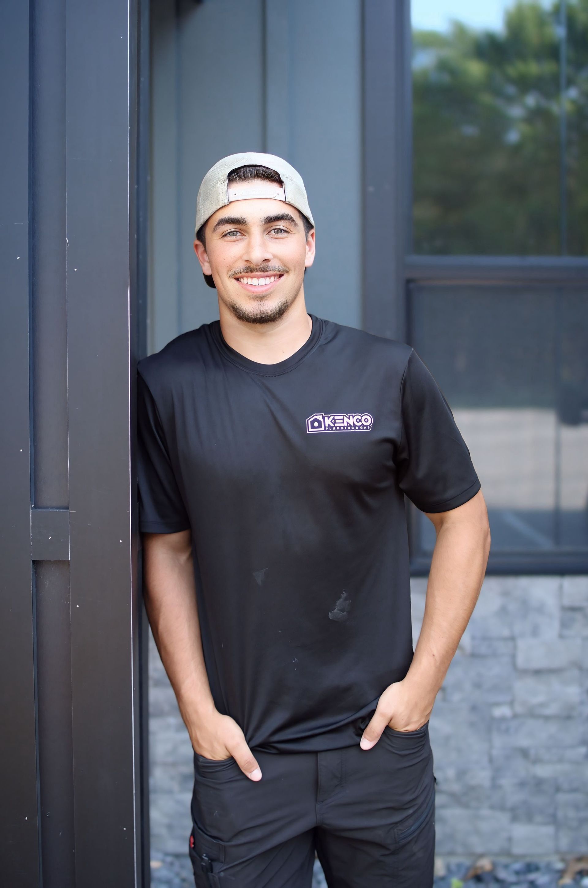 Man in a cap and black shirt, leaning against a dark structure, hands in pockets, smiling.