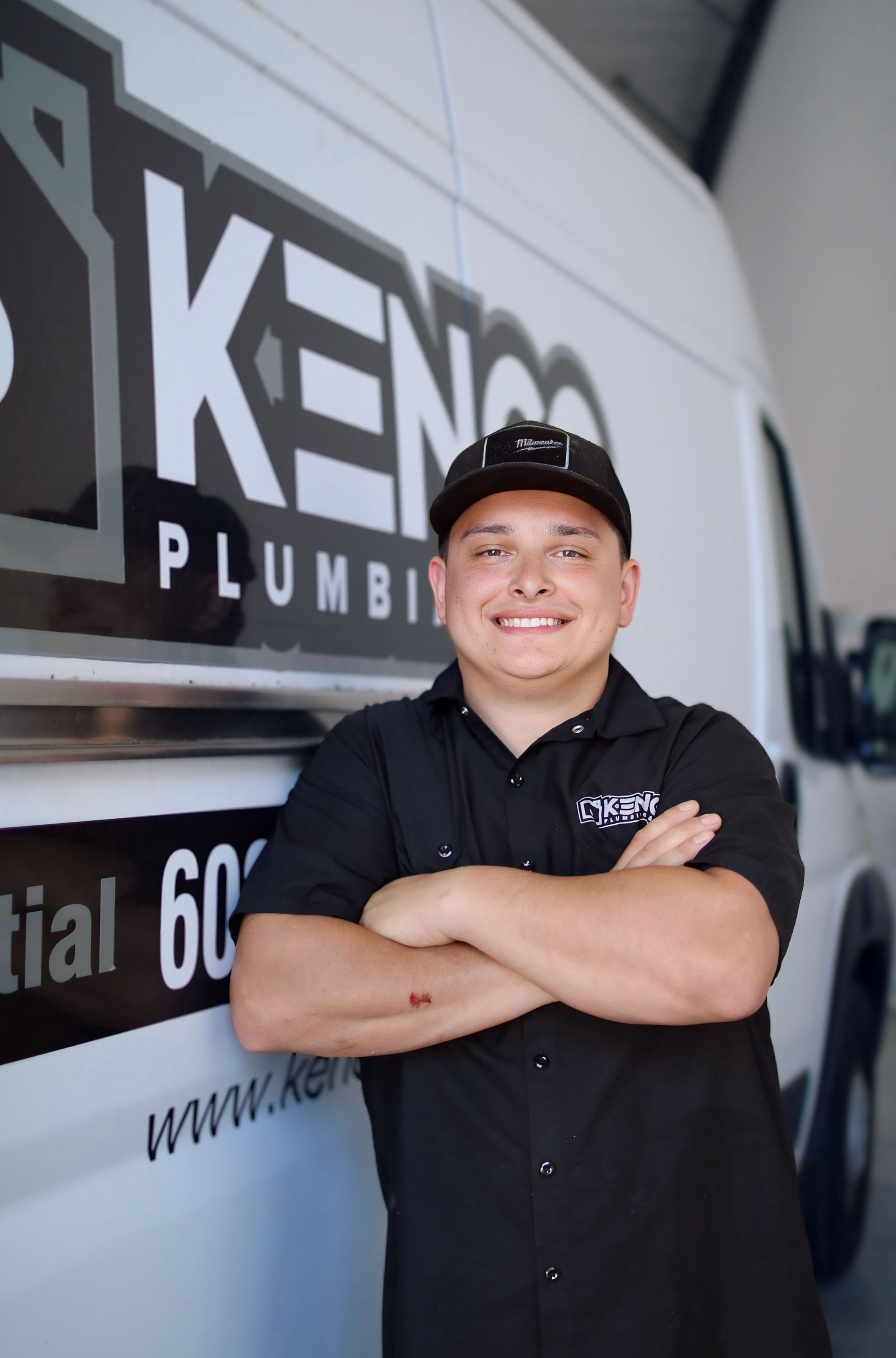 Smiling plumber in black shirt and cap, arms crossed, stands by a Kenko Plumbing van.