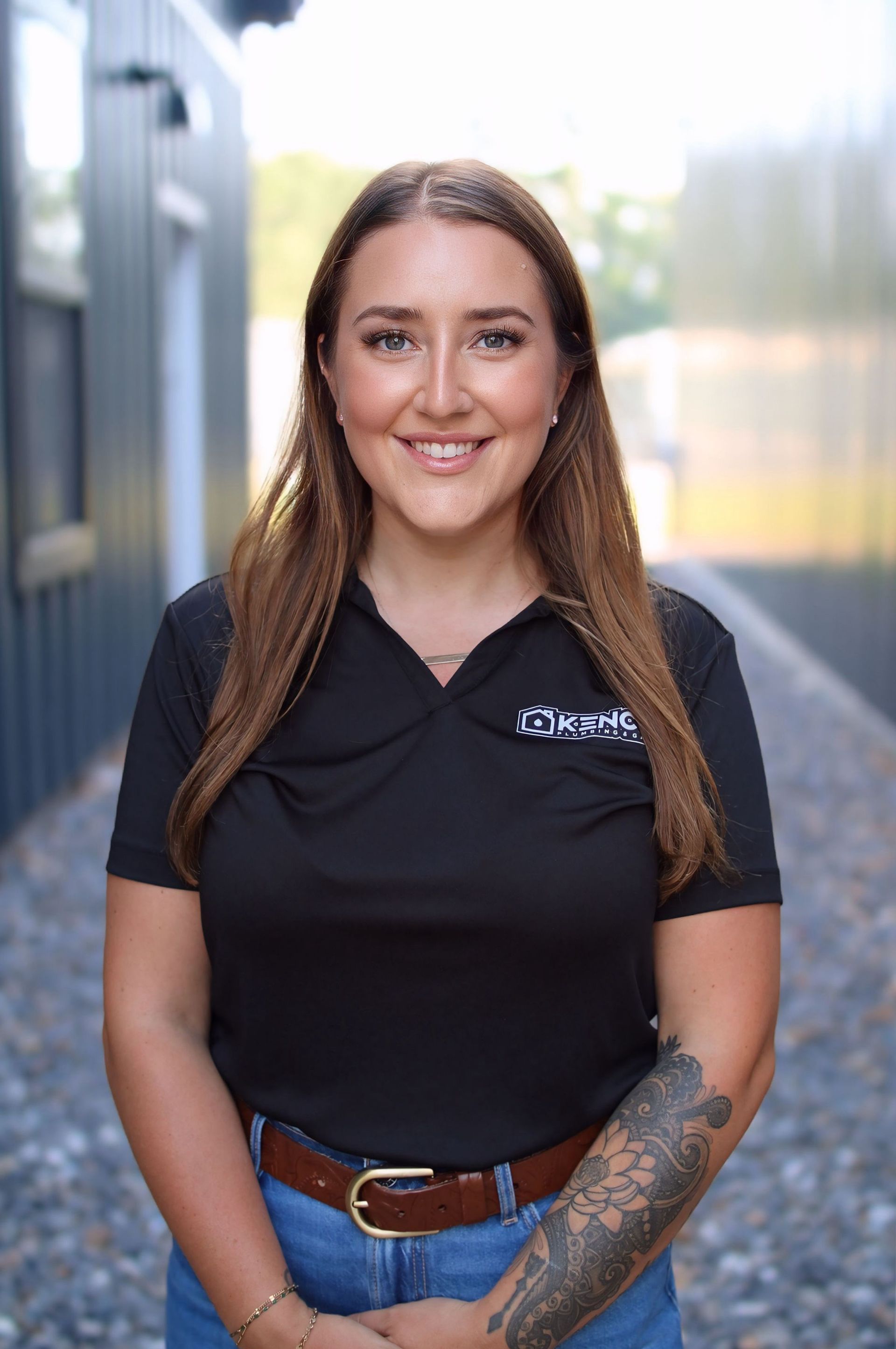 Woman with brown hair, black polo shirt, and arm tattoo, smiling outside.