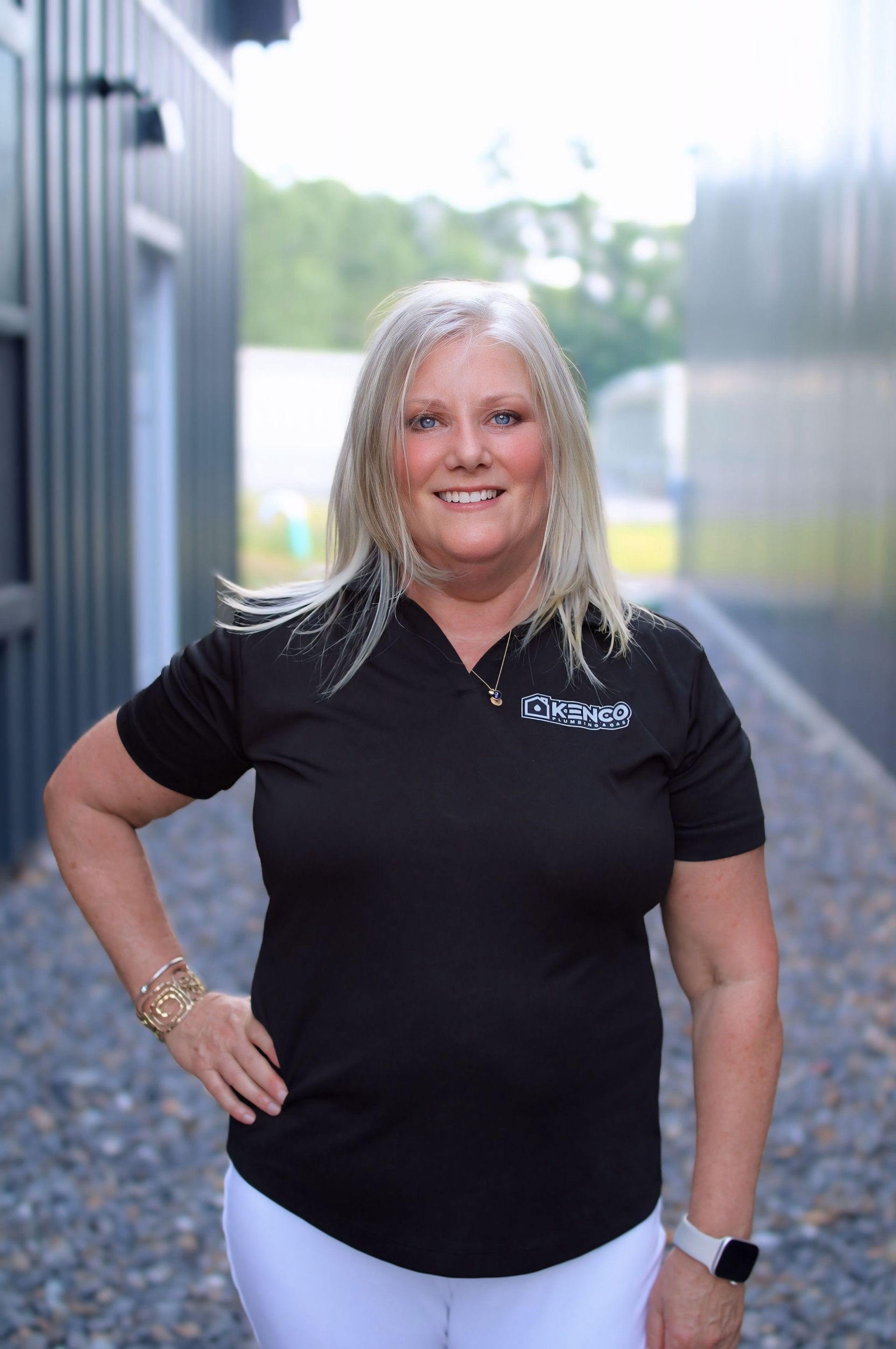 Woman with blonde hair, wearing black polo and white pants, poses with hand on hip outside.